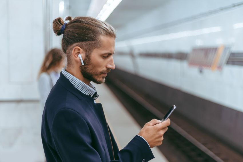 Geschäftsmann steht an einem Bahngleis mit einem Smartphone in der rechten Hand und weißen Bluetooth In-Ear Kopfhörern